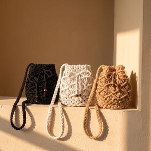 May include: Three bucket bags crafted from knotted cord in black, off-white, and tan. Each bag features a drawstring closure with wooden beads at the ends and a braided shoulder strap. The macrame design creates an open, repeating pattern.