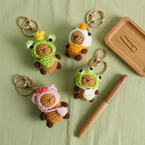 May include: Four crocheted capybara keychains featuring brown yarn bodies. One wears a green frog hood with a yellow crown, one a white chicken hood, one a pink bear hood with a red flower, and one a green frog hood with a white daisy.