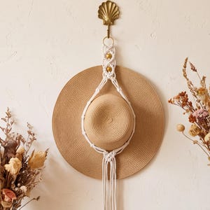 May include: A tan straw hat held by a white macrame cord hanger with two gold beads. The hanger is suspended from a gold-coloured metal wall hook shaped like a seashell. The hat features a wide brim and a rounded crown, hanging vertically.