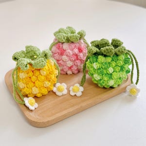 May include: Three small, round, crocheted drawstring pouches shaped like berries. One is yellow, one is pink, and one is green. Each pouch features a green crocheted leaf top, a drawstring closure, and a small white flower attached to the cord ends.