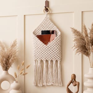 May include: A cream-colored macrame wall hanging organizer featuring a diamond-patterned weave, a circular wooden ring at the top for suspension, and a series of long, knotted fringe tassels at the bottom, containing two stacked notebooks.