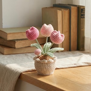 Puede incluir: Un arreglo floral de ganchillo con tres tulipanes rosas y un pequeño capullo en una maceta beige. Las flores hechas a mano tienen tallos y hojas verdes. La maceta tiene un borde festoneado. El fondo incluye libros antiguos y una superficie de madera.