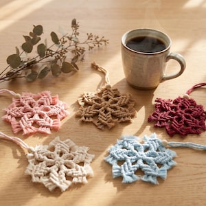 May include: Five macrame snowflake ornaments in pink, beige, dark red, cream, and light blue, each featuring a small wooden bead and a hanging cord. A ceramic mug filled with a dark steaming beverage sits nearby, alongside a sprig of dried eucalyptus.