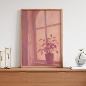 Könnte beinhalten: Ein gerahmter vertikaler Wandkunstdruck mit einem Ölgemälde einer Topfpflanze auf einem Fensterbrett. Die Szene ist in warmen Pfirsich-, Rosa- und sanften Orangetönen gehalten und zeigt das Licht, das durch eine Fensterscheibe auf die Pflanze fällt.