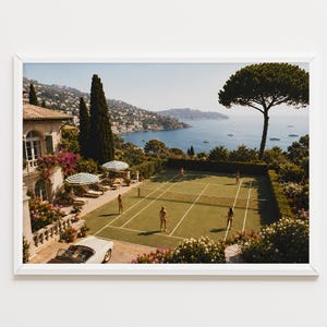 Puede incluir: Una fotografía paisajística enmarcada que muestra una pista de tenis verde con cuatro personas jugando, situada junto a una villa de piedra con un coche descapotable blanco, con vistas a una bahía costera con barcos y colinas bajo el cielo.