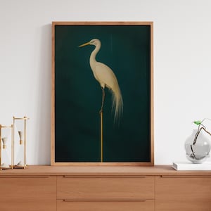 May include: A framed art print of a white egret on a golden rod against a dark teal backdrop. The bird has a long neck and flowing tail feathers. The frame is light-coloured wood. The artwork is displayed on a wooden cabinet. The image is a decorative piece.