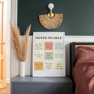 Può includere: Una stampa incorniciata con il testo "NOTES TO SELF" e citazioni motivazionali su post-it colorati. La stampa è su un comodino grigio con un vaso di erba della pampa essiccata. Una lampada da parete decorativa e un letto con una trapunta a motivi rossi sono sullo sfondo.