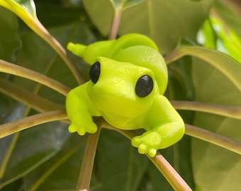 May include: A bright neon yellow frog figurine with a smooth, layered texture and large, round, solid black eyes. The frog is positioned with its limbs extended, resting on the stems of a green plant with broad, glossy leaves.