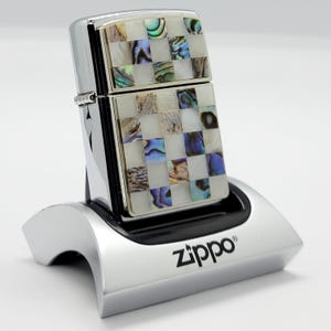 Könnte beinhalten: Ein verchromtes Zippo-Feuerzeug mit einem Schachbrettmuster aus weißen Quadraten und quadratischen Feldern aus schillerndem Abalone-Muschel. Das Feuerzeug steht in einem silbernen Zippo-Markenständer.