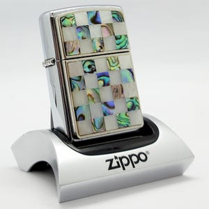 Könnte beinhalten: Ein chromfarbenes Zippo-Feuerzeug mit einem Schachbrettmuster aus schillernden Abalone-Muschelplättchen und weißen Perlmutt-Fliesen auf der Vorderseite. Das Feuerzeug steht aufrecht in einem silberfarbenen Zippo-Kunststoff-Präsentationsständer.