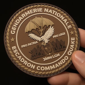 Può includere: Una toppa circolare con le parole "Gendarmerie Nationale" e "Escadron Commando Quake" in un carattere beige. La toppa presenta un paracadute, un'aquila e sagome nei colori marrone e beige.