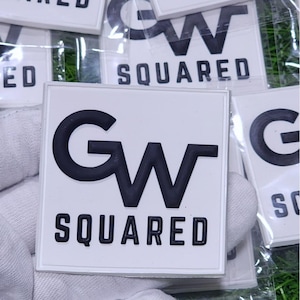 May include: Square white patches with the text "GW SQUARED" in bold black letters. The patches are in a clear plastic bag. The patches are likely for clothing or accessories.