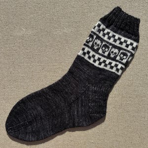 Könnte beinhalten: Ein anthrazitfarbener Strickstrumpf mit geripptem Bündchen und einem dekorativen Band am oberen Teil. Das Band zeigt ein sich wiederholendes Muster aus weißen Totenköpfen, die sich mit kleinen schwarz-weißen Karos auf hellgrauem Grund abwechseln.