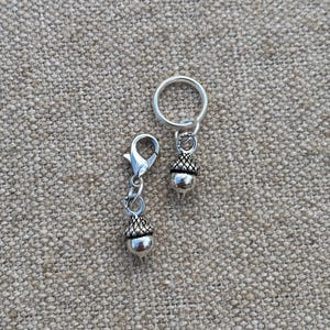 May include: Two silver-colored acorn charms. One charm is attached to a keyring, and the other is attached to a clasp. The acorns have a textured cap and a smooth, rounded body. The background is a textured, tan fabric.