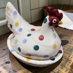 May include: A ceramic chicken-shaped egg holder with a white base featuring multi-coloured polka dots in red, blue, yellow, and green. The chicken has a dark red comb, a yellow beak, and a white eye with a black pupil, resting on a matching dish.