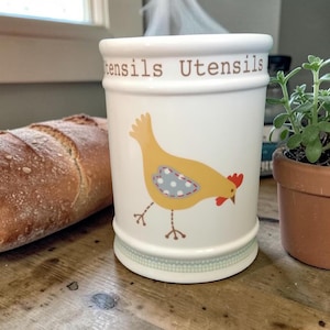 May include: A white ceramic utensil holder featuring a yellow chicken illustration with a blue and white polka dot wing patch. The word Utensils is printed in brown text near the top rim, and a light blue patterned band encircles the base.
