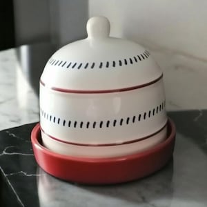 May include: A white ceramic butter dish with a red base and lid. The lid has a small knob and is decorated with black dashes and a red band. The dish sits on a red plate, creating a contrasting colour scheme.