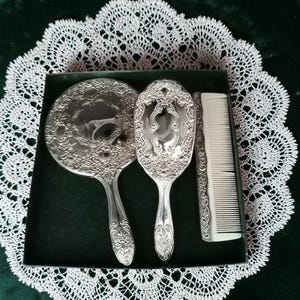 May include: A vintage vanity set comprising a hand mirror, brush, and comb, featuring ornate silver-toned floral patterns. The set is displayed in a black box, set upon a white lace doily, against a dark green backdrop.