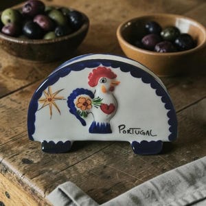 May include: A semi-circular ceramic napkin holder with a white base and a dark blue scalloped border. It features a hand-painted rooster with a red comb, a yellow flower, and a star. The word PORTUGAL is written in black script on the front.