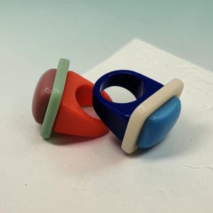 May include: Two chunky resin rings with geometric designs. One ring features a bright orange band with a muted red dome and a pale green rectangular accent. The second ring has a deep blue band with a light blue dome and a cream rectangular accent.