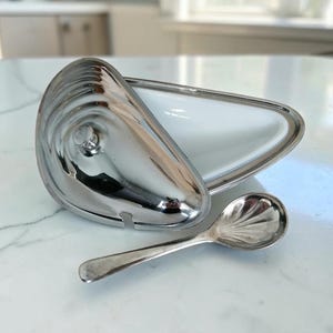 May include: A silver-toned metal serving dish shaped like a mussel shell with a hinged lid and a white interior, accompanied by a small silver-toned metal spoon featuring a shell-patterned bowl, both resting on a white surface.