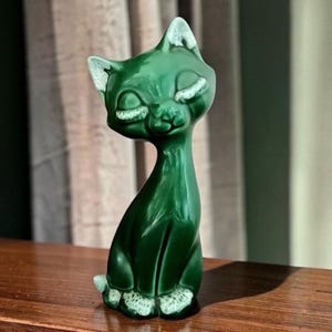 May include: A glossy dark green ceramic cat figurine with an elongated neck and closed eyes. The cat features white textured accents on the tips of its ears, eyelids, and paws, creating a distinct contrast against the smooth, deep green glazed finish.