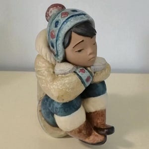 May include: A ceramic figurine of a person in winter clothing, seated with knees drawn up. The figure wears a light blue hat with red and blue details, a cream-coloured parka, and blue trousers. Brown boots and mittens complete the outfit. The figurine has a thoughtful expression.