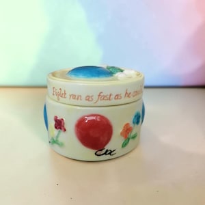 May include: A round, cream-coloured ceramic trinket box with a lid. The lid features a blue accent and the text "Piglet ran as fast as he could". The base is decorated with a large red circle, small colourful flowers, and the black signature "Cox".