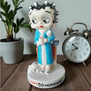 May include: A ceramic figurine of a cartoon character with black hair, wearing a blue robe and white slippers, holding a white mug. The character stands on a round white base with the text I DON'T DO MORNINGS! printed in black and red letters.