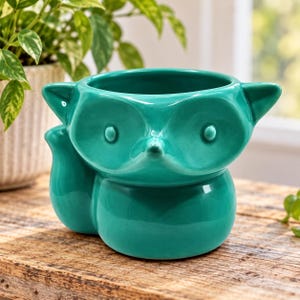 May include: A teal ceramic planter shaped like a fox head with pointed ears and a curved tail on the side. The smooth, glossy surface features a stylised face with circular eyes and a small, pointed nose, creating a hollow vessel for plants.