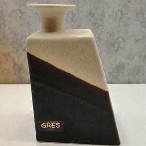 May include: A ceramic vase with an angled design. The upper part is beige, transitioning to a dark brown diagonal section. The vase has a flared top and a label that reads "GRES 1300 C".