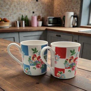 May include: Two white ceramic mugs with floral designs and checkered patterns in blue and red. The mugs have curved handles and are set on a wooden surface, suggesting a kitchen setting.