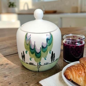 May include: A white ceramic sugar bowl with a lid featuring a rounded knob handle. The exterior is decorated with a hand-painted landscape scene in shades of green, teal, and lavender, depicting stylised trees and a grassy horizon line.