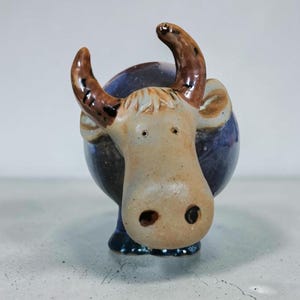 May include: A ceramic figurine of a bull with a round, dark blue body and a light tan head. The bull features two curved, brown-spotted horns, small eyes, and a wide snout with two dark nostrils. The piece has a smooth, glazed finish throughout.