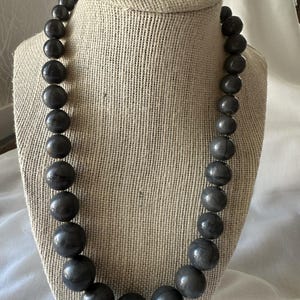 Op de afbeelding: Een enkelstrengs ketting met gegradueerde ronde kralen in een donker antracietgrijze kleur. De kralen nemen in grootte toe vanaf de sluiting naar het midden, waar de grootste kraal zich onderaan de cirkelvormige opstelling bevindt.