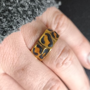 Könnte beinhalten: Ein goldfarbener Ring mit Leopardenmuster. Der Ring hat schwarze Flecken auf goldenem Hintergrund. Der Ring wird an einem Finger getragen. Der Ring ist ein Modeaccessoire.