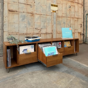 May include: A wooden record player stand with a drawer that is pulled out. The stand has multiple shelves for storing vinyl records. The stand is in front of a metal garage door with a "High Voltage" sign. There are several vinyl records visible in the shelves and drawer, including "I Was Real", "Paradisa", and "Joan Bibiloni".