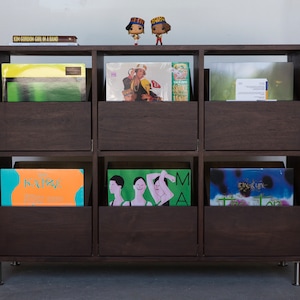 A 6-drawer cabinet in a dark brown stain with LPs stored inside the drawers. There are two books and two figurines on the top of the cabinet. The legs are chrome.