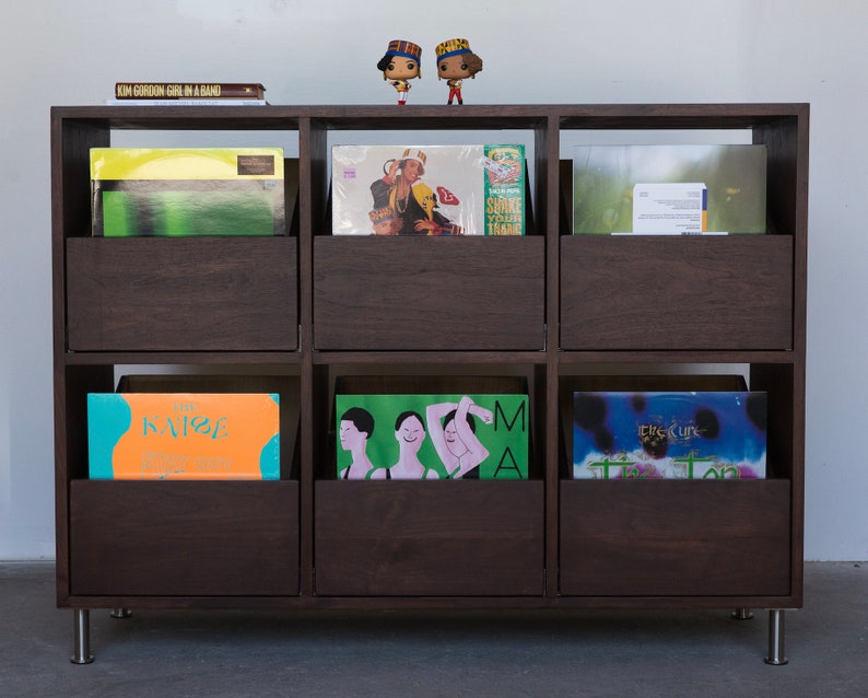 A 6-drawer cabinet in a dark brown stain with LPs stored inside the drawers. There are two books and two figurines on the top of the cabinet. The legs are chrome.