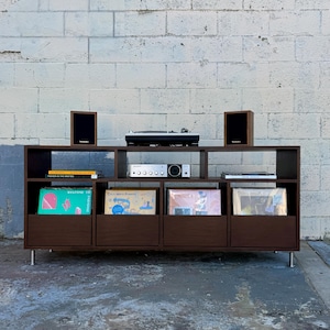 May include: A dark brown wooden record player stand with six shelves. The stand has a turntable, a stereo receiver, and two speakers. There are vinyl records on the shelves. The stand is in front of a white brick wall.