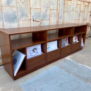 May include: A long, rectangular wooden record storage unit with multiple compartments. The unit is made of a warm-toned wood and features open shelves and drawers, with vinyl records visible in some compartments. The unit is placed against a textured wall.