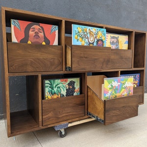 May include: A walnut wood record player stand with multiple shelves and a drawer. The stand has a variety of vinyl records with colorful artwork displayed on the shelves.