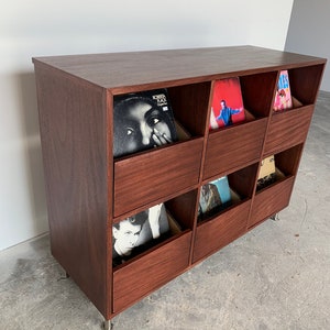 A side view of a six-drawer record storage cabinet in a medium brown stain with vinyl records inside the drawers. The legs are chrome.