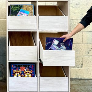 May include: A light-colored wooden storage unit with six open compartments and a pull-out drawer. The unit features a light wood grain finish and silver metal legs. Several colorful rectangular items are stored inside the various compartments.