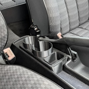 以下が含まれることがあります： 2つの円形カップホルダーと長方形の収納コンパートメントを備えた、黒いプラスチック製の車のコンソールカップホルダー。コンソールは、模様のある灰色の生地の2つのカーシートの間に配置されています。「PRESS」という文字が入ったシートベルトバックルが見えます。