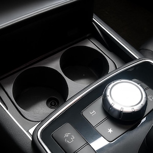 Puede incluir: Primer plano del interior de un coche, que muestra una consola negra con un portavasos y un panel de control. El panel de control presenta un dial plateado y varios botones. El portavasos tiene dos compartimentos circulares.