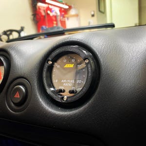 Peut inclure: Un indicateur de rapport air/carburant AEM noir installé dans le tableau de bord d'un véhicule. L'indicateur affiche des chiffres de 8 à 20, avec le logo AEM et le texte "AIR/FUEL RATIO". La jauge est entourée d'une lunette noire et fixée avec des vis.
