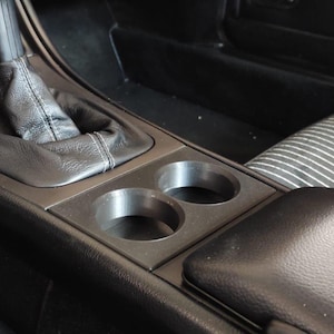 May include: Interior car console with a gear shift, two cup holders, and an armrest. The console is black with a leather-like texture. The seats have a striped pattern.