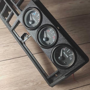 Può includere: Un pannello cruscotto nero con tre indicatori rotondi. Gli indicatori sono etichettati "TEMP C", "VOLT" e "OIL KG".