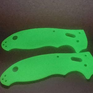 Op de afbeelding: Twee groene, in het donker lichtgevende mes schalen. De schalen zijn gemaakt van een glad, plastic-achtig materiaal en hebben een licht gebogen vorm.